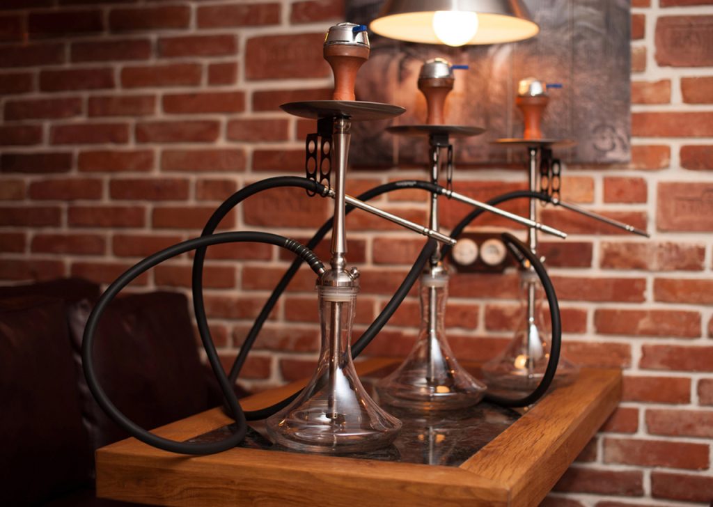 Premium stainless steel hookahs with clear glass bases and black hoses set up on a table against brick wall in Houston's best hookah shop.