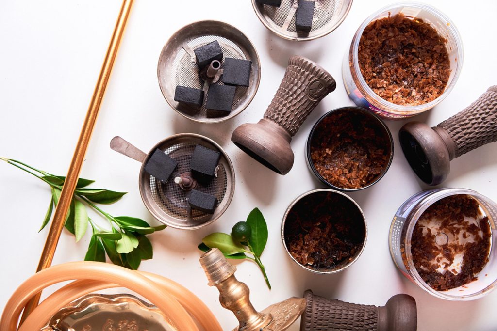 Variety of premium shisha tobacco flavors in jars, hookah bowls with coals, and accessories displayed at a leading tobacco shop in Houston.