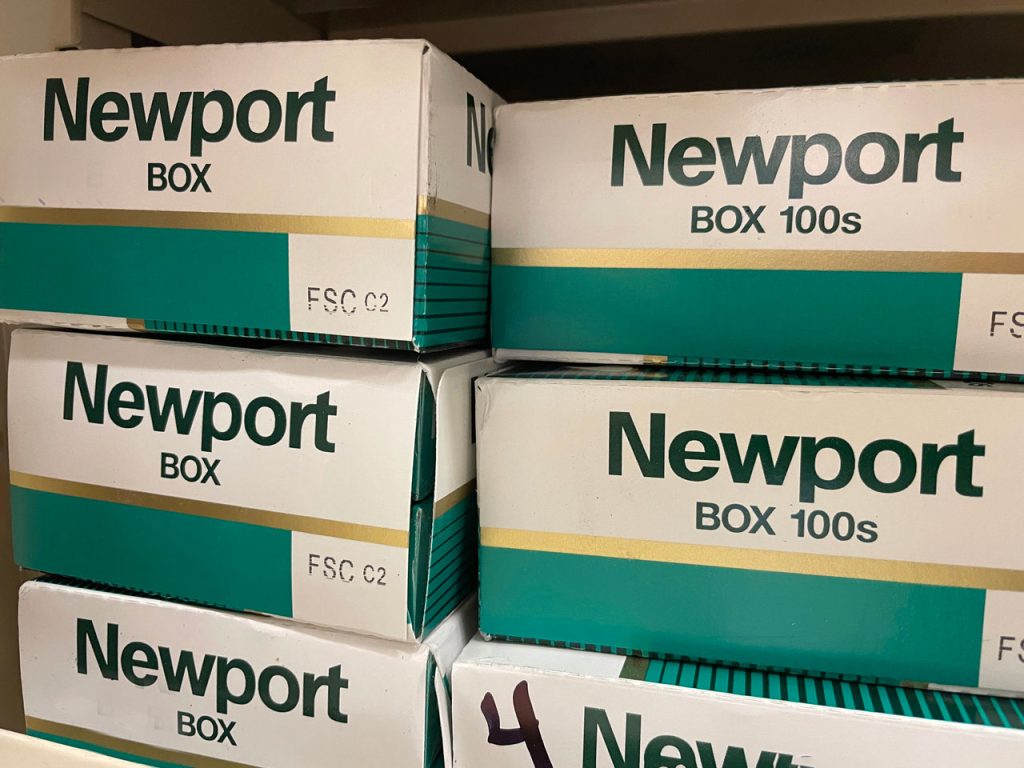 A close-up of stacked Newport cigarette cartons, including Box and Box 100s sizes, on a retail shelf display at a cigarette shop Houston.