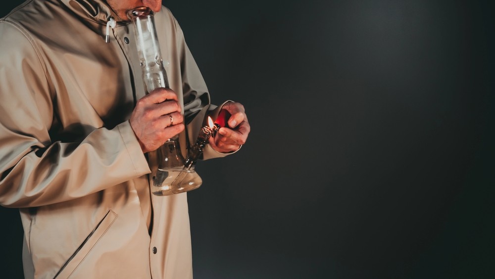 A person in a beige jacket uses a lighter on a clear glass water pipe, a common product at a head shop Houston. 
