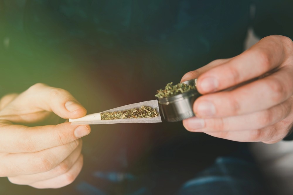 Close up of hands pouring ground herbs from a metal grinder into a rolling paper from a head shop Houston supply. 
