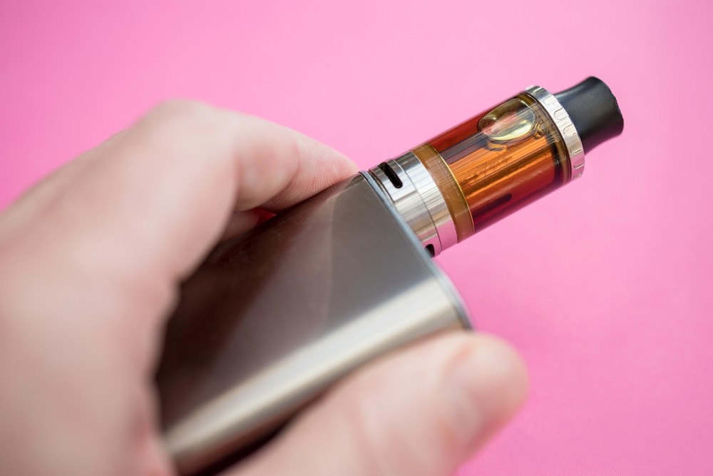 Close-up of a hand adjusting a silver vape pen filled with amber e-liquid on a vibrant pink background, available at top smoke shop Houston for premium vaping experiences.