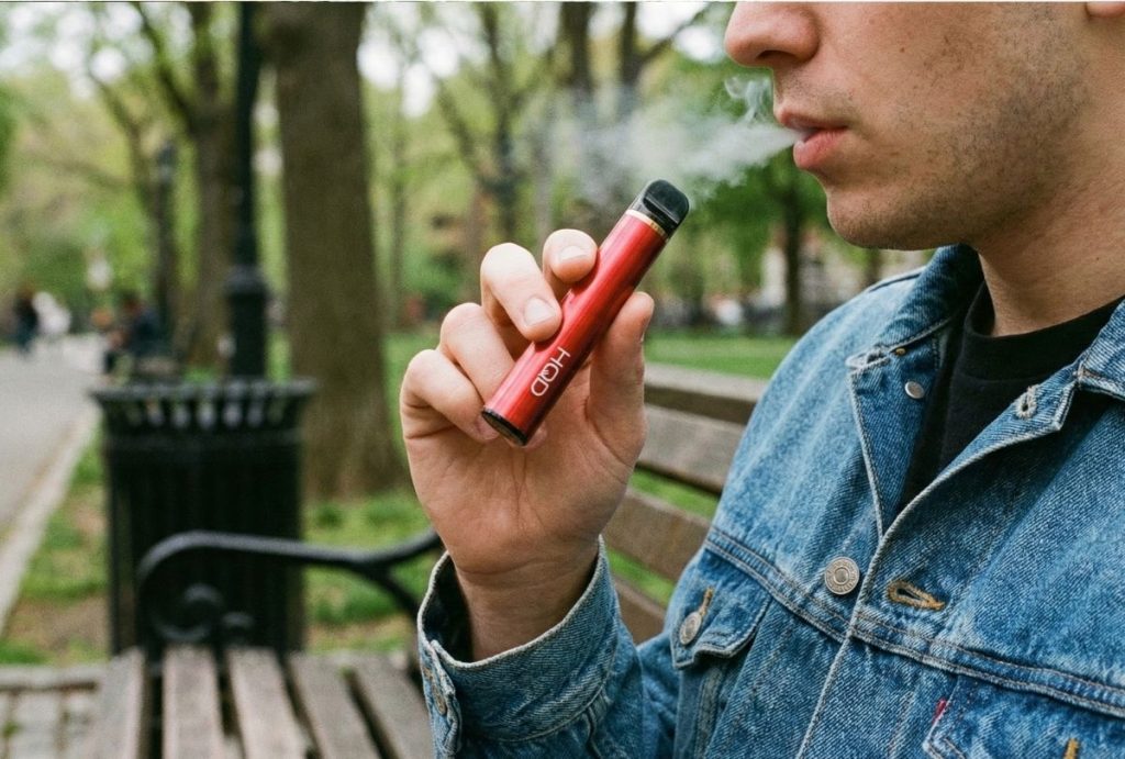 Man exhaling vapor from a red disposable HQD vape in a park, a habit that might require an Ambetter urgent care visit.