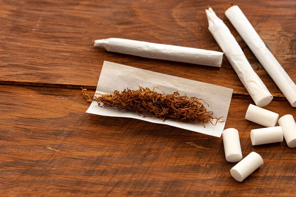 Pre-rolled cigarettes and white filters surround loose tobacco ready for rolling papers houston on a rustic wood surface.