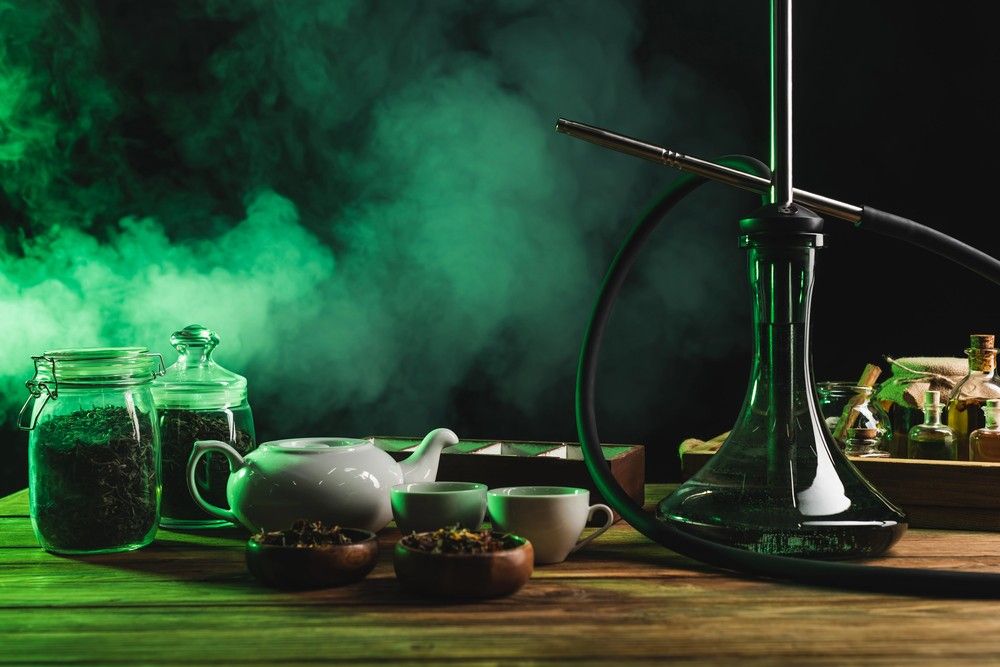 A glass shisha pipe sits on a wooden table next to a white teapot, showcasing items found at a head shop Houston.